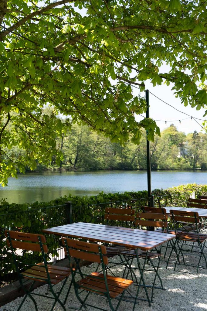 Biergarten am See mit Holztischen, Stühlen und schattigem Außenbereich unter Bäumen.