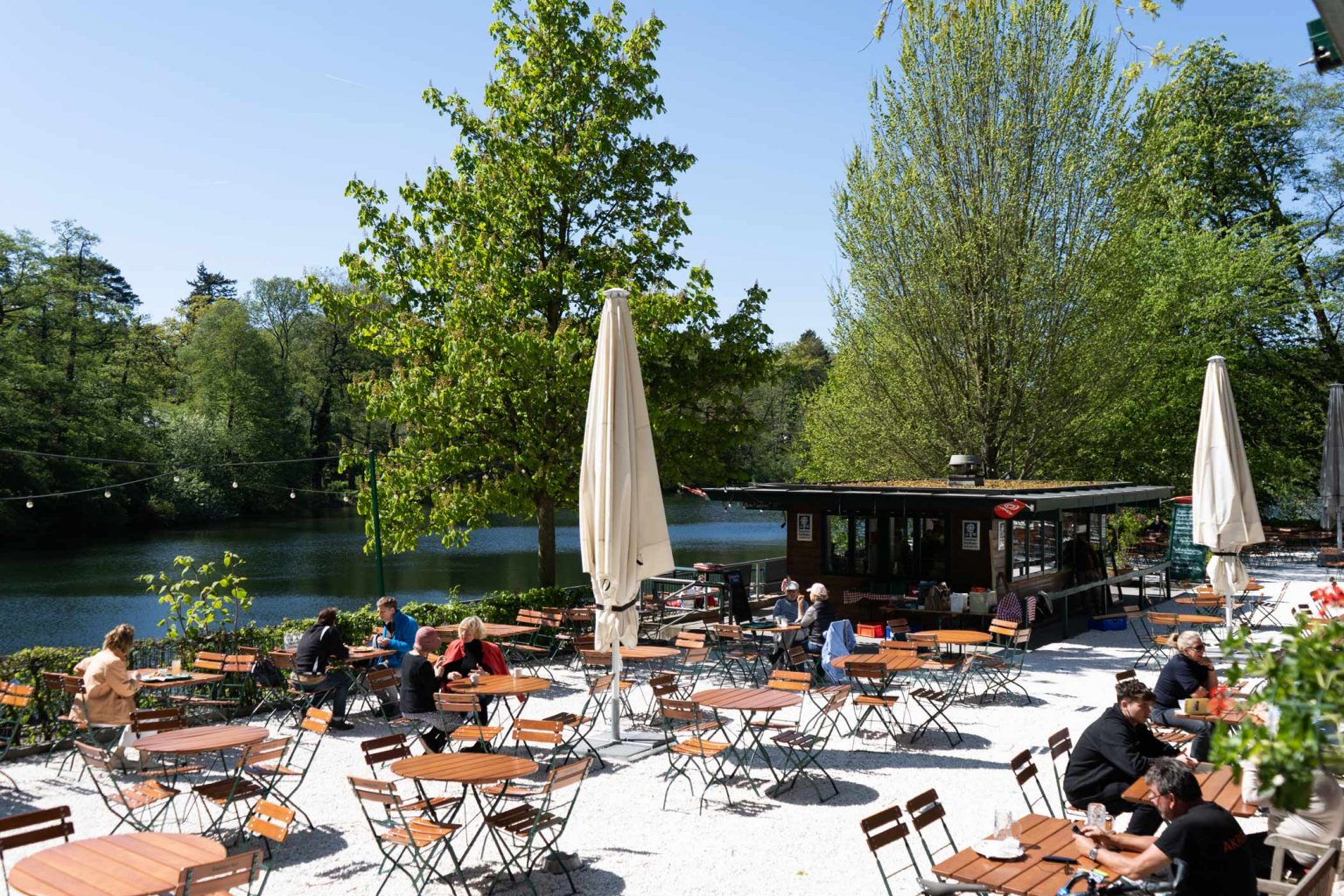 Biergarten am See mit Sonnenschirmen, Holztischen und grünem Außenbereich.