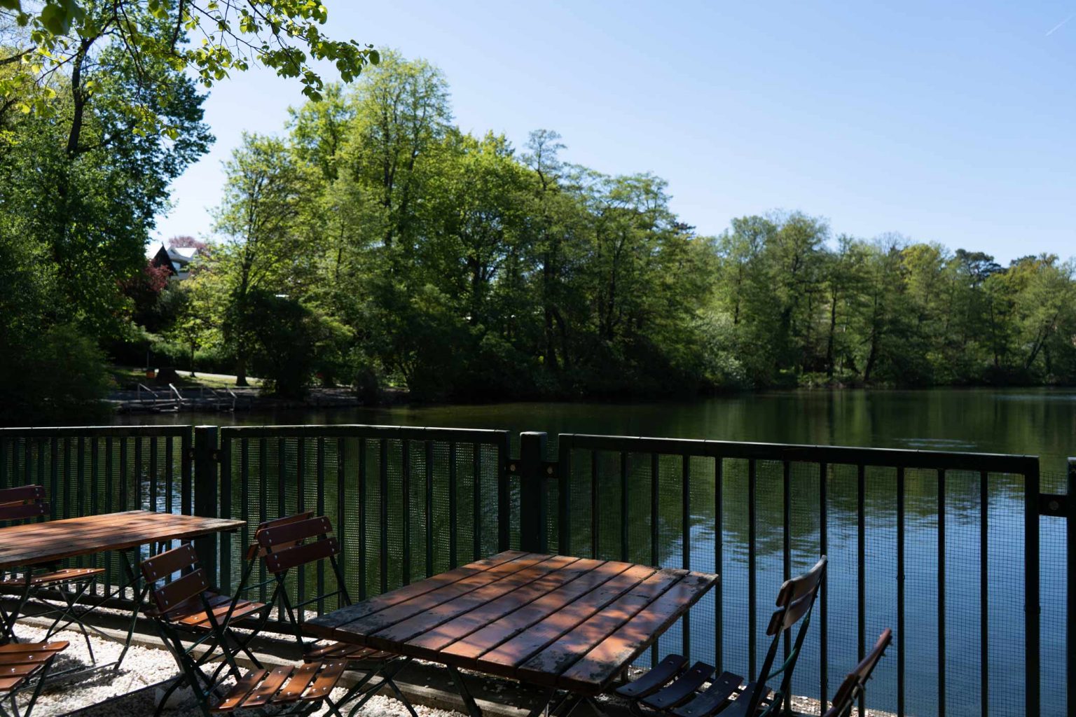 Biergarten-Sitzplätze am Geländer mit Blick auf den See und grünes Ufer.