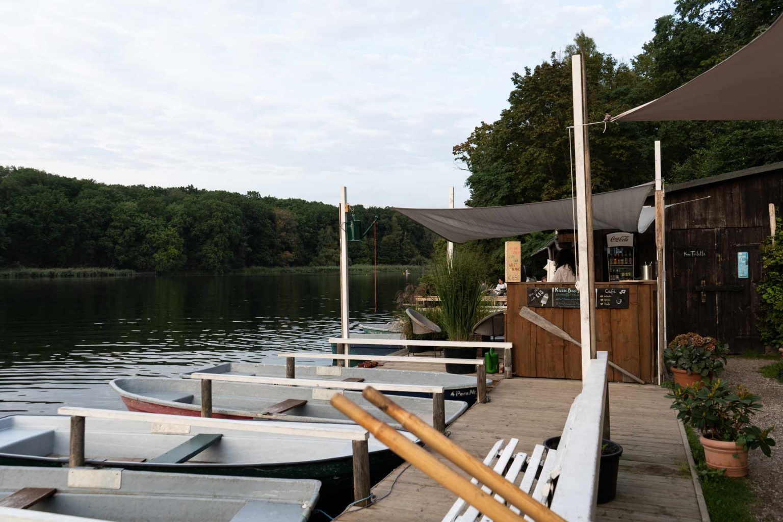 Ruderboote am Seeufer mit Bootssteg, Kiosk und Wald im Hintergrund.