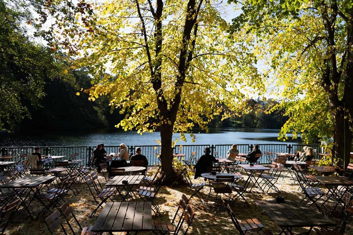 Biergarten unter Bäumen mit Holztischen, Stühlen und direktem Blick auf den See.