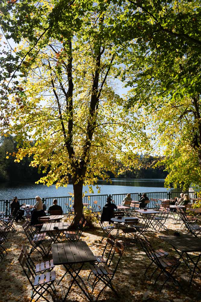 Biergarten am See mit Holztischen, Sitzplätzen unter Bäumen und herbstlichem Blick aufs Wasser.