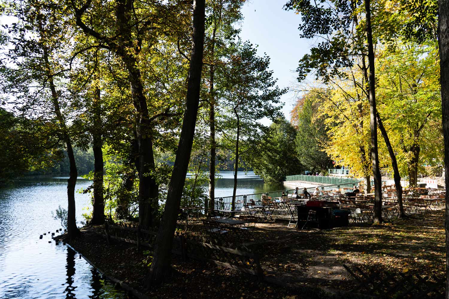 Ruhiger Biergarten am Seeufer mit Sitzplätzen unter Bäumen und Blick aufs Wasser.